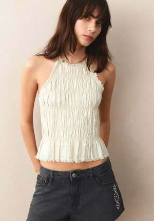 Jeune femme aux longs cheveux foncés portant un top blanc sans manches froncé et un short en jean noir avec des clous décoratifs.