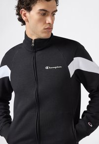 Uomo che indossa una giacca Champion nera con zip full-length, con strisce bianche sulle maniche e logo sul petto, mentre guarda verso sinistra.