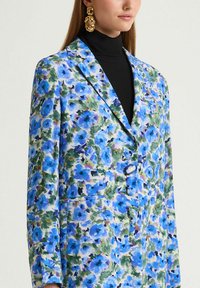 Blazer floral en tissu avec des fleurs bleues et violettes sur un fond clair, doté d'un bouton unique et d'un col à revers cranté.