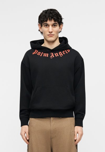 Jeune homme aux cheveux courts et bouclés portant un sweat à capuche noir avec le texte rouge "Palm Angels" et un pantalon beige, sur fond uni.