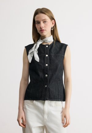 Femme portant un haut noir sans manches boutonné, une écharpe blanche à motifs nouée autour du cou, et un pantalon blanc, debout devant un fond uni.