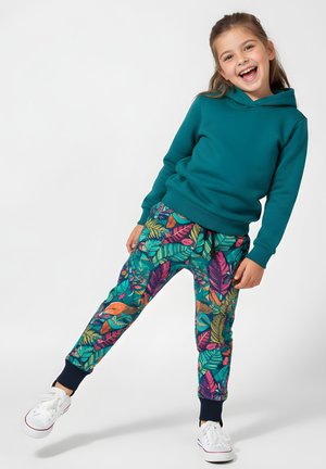 Uśmiechnięta dziewczyna w turkusowej bluzie z kapturem, jaskrawych spodniach w tropikalny wzór liści i białych trampkach stoi na jednej nodze na jednolitym tle.