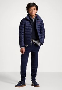 Gutt som står iført en marineblå pufferjakke, stripete skjorte, marineblå genser, joggebukser og marineblå sneakers med gummisåler mot en enkel hvit bakgrunn.