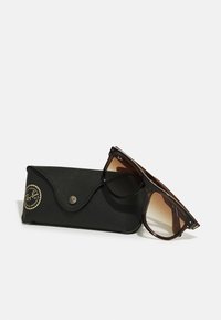 Ochelari de soare aviator cu un gradient maro, într-un toc din piele neagră cu un logo aurit. Ochelarii au rame groase și un finisaj lucios.