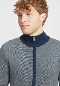 Suéter de punto azul marino y blanco con cremallera, cuello acanalado y patrón diagonal. Cuenta con una cremallera frontal y una tela texturizada.