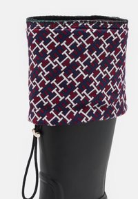 Botte en caoutchouc noire avec un revers en tissu maroon et blanc à motif, comportant un cordon de serrage et des éléments décoratifs.