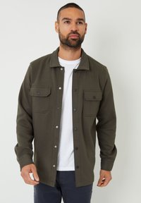 Chaqueta de camisa color verde oliva con botones, dos bolsillos frontales, un cuello y una textura suave. Llevada sobre una camiseta blanca y pantalones oscuros.