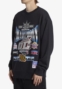 Black sweatshirt with crew neck, featuring large graphic designs and various logos in silver, red, and blue. Notable patches present.