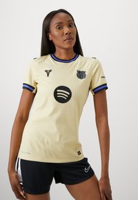 Ljusgul kortärmad tröja i texturerad tyg, med svart Spotify-logotyp och FC Barcelona-emblem, accentuerad av lila och svart kant.