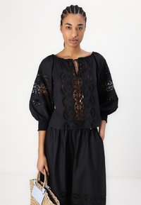 Blusa nera con ricami, maniche a sbuffo e scollatura con cordoncino. Indossata con una gonna nera coordinata. Il modello tiene una borsa in tessuto.