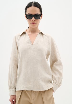 Beige linen top with a V-neck, long sleeves, and gathered cuffs. The fabric has a natural texture, paired with tan trousers and dark sunglasses.