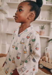 Chemise de pyjama blanche avec col, ornée de motifs de lapins colorés. Tissu doux avec fermeture à boutons et design froncé à la taille.