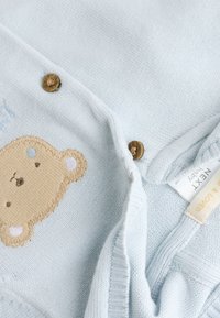Cardigan bébé bleu clair avec un patch d'ours brun sur le devant et des boutons en bois, montrant les étiquettes de vêtements à l'intérieur du col.