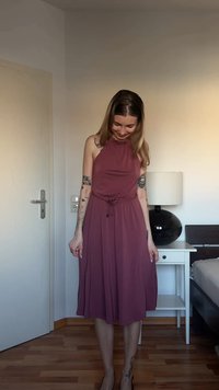 Γυναίκα με μακριά μαλλιά που φοράει αμάνικο φόρεμα σε αποχρώσεις του μωβ, στέκεται σε ένα υπνοδωμάτιο δίπλα σε ένα λευκό κομοδίνο με μαύρο φωτιστικό.