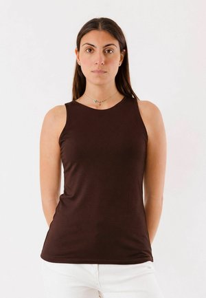 Donna con capelli lunghi e scuri, indossa una canotta marrone scuro e pantaloni bianchi, in piedi davanti a uno sfondo chiaro e uniforme.