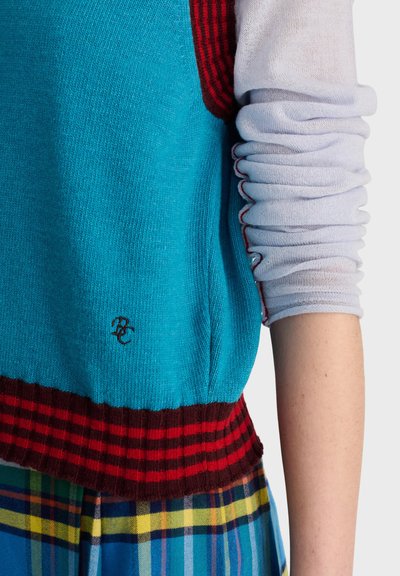 Bras portant une chemise bleu clair à manches longues retroussées sous un gilet en tricot bleu avec des bordures rouges et marron sur une jupe à carreaux multicolores.
