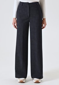 Pantaloni a gamba ampia in tessuto blu scuro con una texture sottile, dotati di chiusura a bottone e zip, e tasche frontali; abbinati a sneakers bianche.