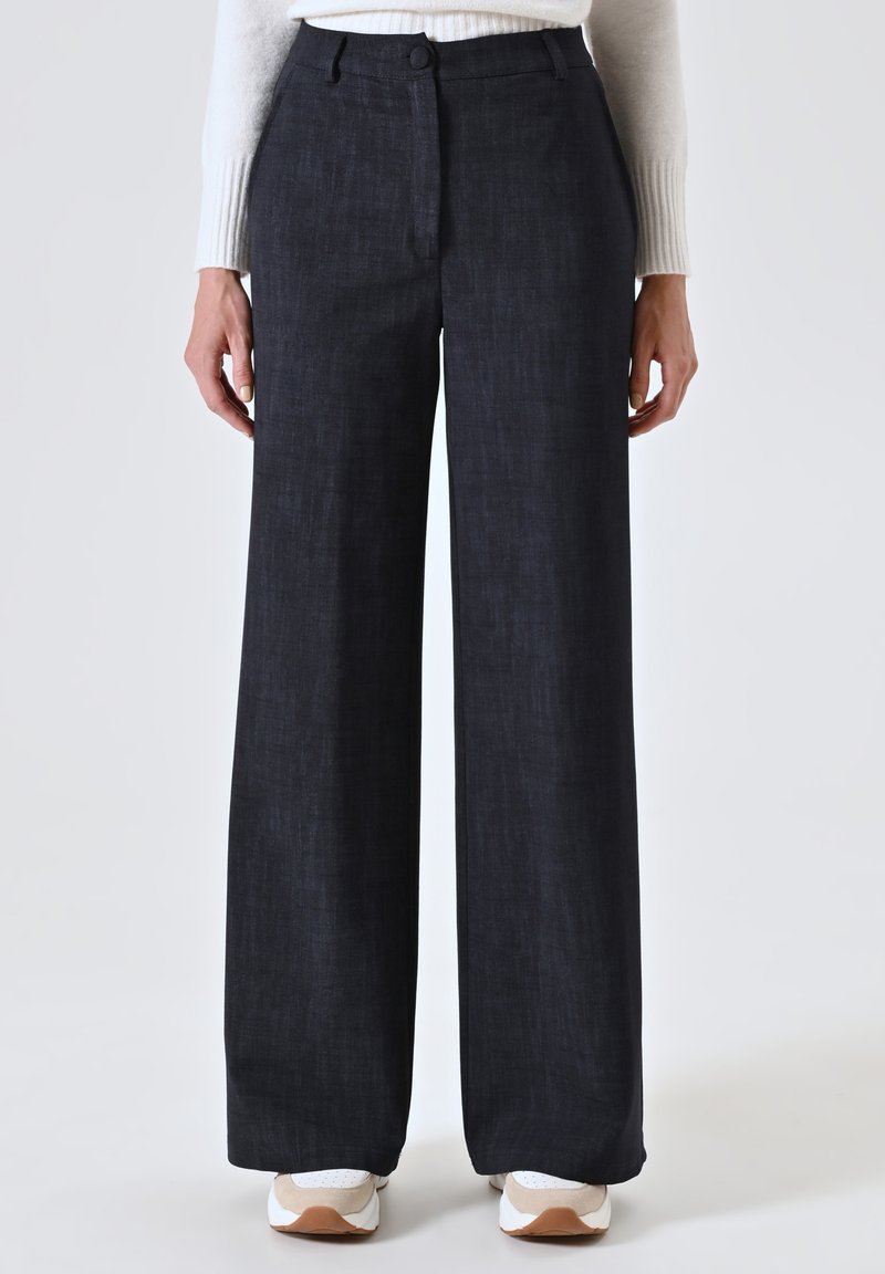 Pantaloni a gamba ampia in tessuto blu scuro con una texture sottile, dotati di chiusura a bottone e zip, e tasche frontali; abbinati a sneakers bianche.