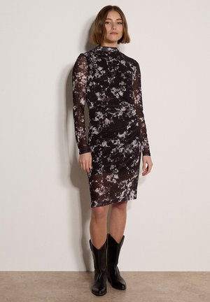 Femme debout contre un mur, portant une robe à manches longues noir et blanc à motifs et des bottes noires mi-mollet avec une coupe au carré.