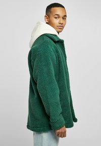 Grønn, fluffy fleecejakke med krage, lange ermer og rund kant. Brukt over en hvit hettegenser.