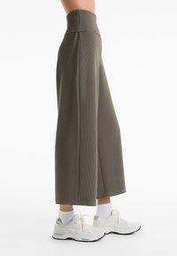 Hoch taillierte, weit geschnittene Hosen aus olivgrünem Rippmaterial. Glatte Textur mit seitlichen Nähten, kombiniert mit weißen Sneakers und Knöchelsocken.