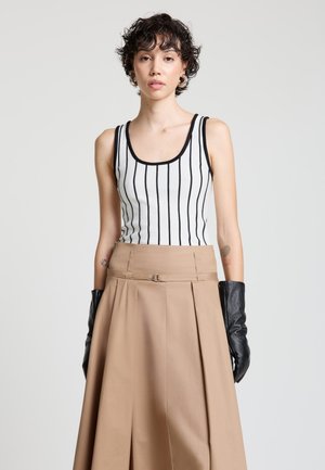 Giovane donna che indossa una canottiera bianca con strisce verticali nere, gonna beige a vita alta, guanti in pelle neri e orecchini a cerchio d'argento.