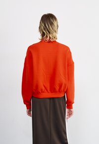 Rød hettegenser med løs passform, ribbestrikket nederkant og senkede skuldre, kombinert med et langt brunt skjørt og minimale detaljer.