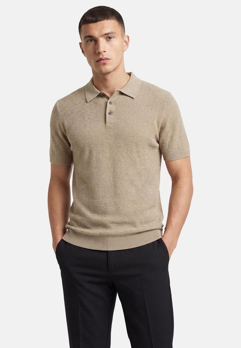 Jeune homme portant un polo en tricot beige à manches courtes avec des boutons, associé à un pantalon noir, les mains dans les poches, sur fond blanc.