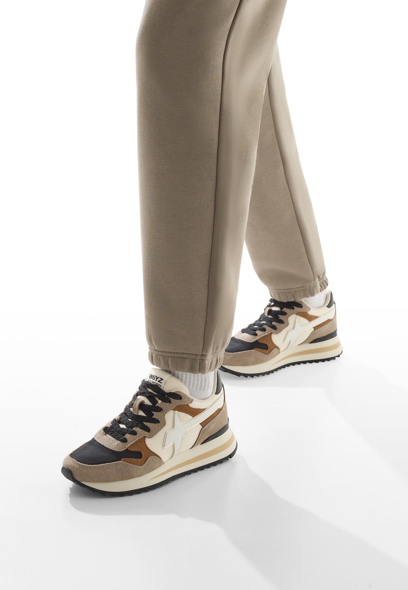 Persona che indossa jogging beige e sneaker beige, nere e bianche con calzini bianchi, in piedi su una superficie bianca.