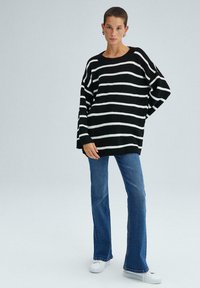 Svart- och vitt randig stickad tröja med vida ärmar, avslappnad passform, ribbat mönster, tillsammans med utsvängda jeans av denim och vita sneakers.