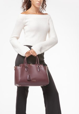 Frau trägt weißen schulterfreien Pullover und schwarze Hose, hält eine burgunderrote Lederhandtasche mit goldenen Beschlägen.