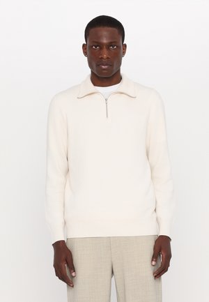 Suéter cremado con cremallera hecho de un tejido suave, que presenta un pequeño cuello y puños acanalados, combinado con pantalones gris claro de corte entallado.