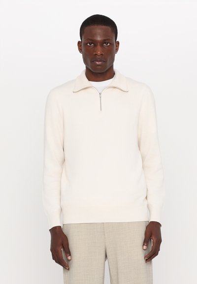 Pull zip beige clair en tissu doux, doté d'un petit col et de poignets côtelés, associé à un pantalon taillé gris clair.