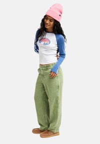 Langärmeliges weißes Hemd mit blauen Ärmeln und grafischem Design, kombiniert mit grünen weit geschnittenen Hosen und braunen Slip-On-Schuhen. Pinke Mütze auf dem Kopf.