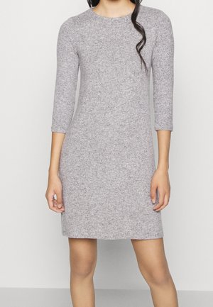 Kvinde iført en knælængde, lysegrå, langærmet strikkjole, stående foran en ensartet hvid baggrund.