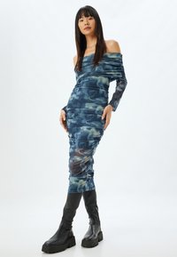 Vestido ajustado y de hombros al aire en patrones abstractos azul y verde, con textura fruncida y mangas largas y transparentes. Botas negras hasta la rodilla.