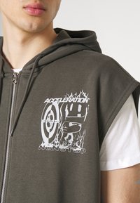 Hombre que lleva una sudadera sin mangas de cremallera gris oscuro con un gráfico blanco que dice "ACELERACIÓN SIN PARAR" sobre una camiseta blanca parcialmente visible.