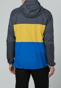Giacca color block con parte superiore a motivo a griglia nera, sezione centrale gialla e sezione inferiore blu vivace. Presenta un cappuccio e un orlo elastico.