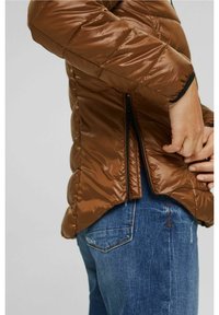 Giacca zip-up marrone, soffice, con finitura lucida, caratterizzata da dettagli neri lungo i polsini e le cerniere laterali, indossata sopra dei jeans blu.