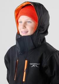 Zwarte weersbestendige jas met een oranje gevoerde capuchon, voorzien van oranje accenten en een logo. Gestructureerde stof, met ritszakken.