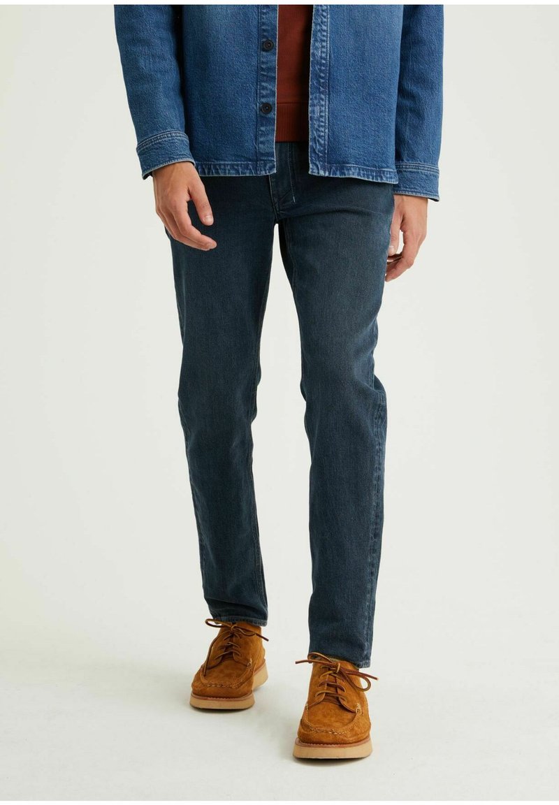 Blauwe denimjas met zichtbare knopen, bruine sweater, donkerblauwe jeans en bruine suède schoenen met veters, die een casual outfit tonen.