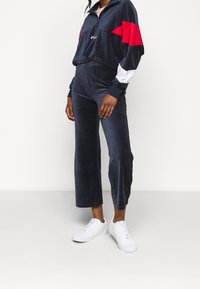 Marinblå velour-träningsdräkt med zippered krage, ljusrosa undertröja och vita accenter. Bärs med vita sneakers. Har byxor med avslappnad passform.