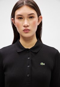 Μαύρο πικέ πόλο μπλουζάκι με καμπύλο γιακά, εμπρόσθιους κουμπιά και μικρό κεντητό λογότυπο κροκόδειλου σε πράσινο χρώμα στην αριστερή πλευρά.