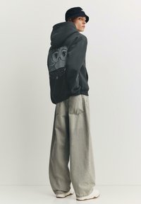 Fekete kapucnis pulóver hátul egy grafikával, világosszürke bő szárú nadrággal és fehér tornacipővel párosítva. A bucket kalap teszi teljessé a megjelenést.