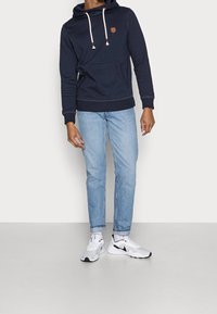 Námořnická mikina s přední kapsou, nastavitelnými šňůrkami a koženou nášivkou s logem. Kombinována se světle modrými džíny a bílými teniskami.