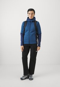 Giacca blu con zip e cappuccio, pantaloni cargo neri e scarpe da trekking grigie. Presenta una consistenza del tessuto liscia e accenti di blu scuro.