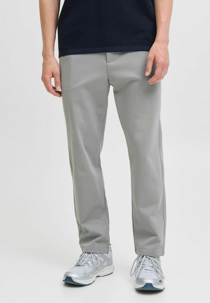 Uomo che indossa pantaloni dritti grigi chiari e scarpe da ginnastica argentate, in piedi con le mani rilassate ai fianchi su uno sfondo bianco uniforme.