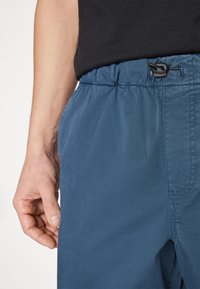 Blue pants made from a lightweight fabric feature an elastic waistband with a drawstring, side pockets, and a smooth texture.
