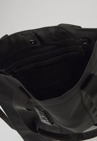 Černá taška přes rameno s textilními a koženými akcenty. Obsahuje vnitřní zipovou kapsu, plochou kapsu a obdélníkový tvar. Viditelný strukturovaný povrch.