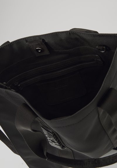 Black tote bag with fabric and leather accents. Features interior zip pocket, flat slip pocket, and rectangular shape. Textured surface visible.